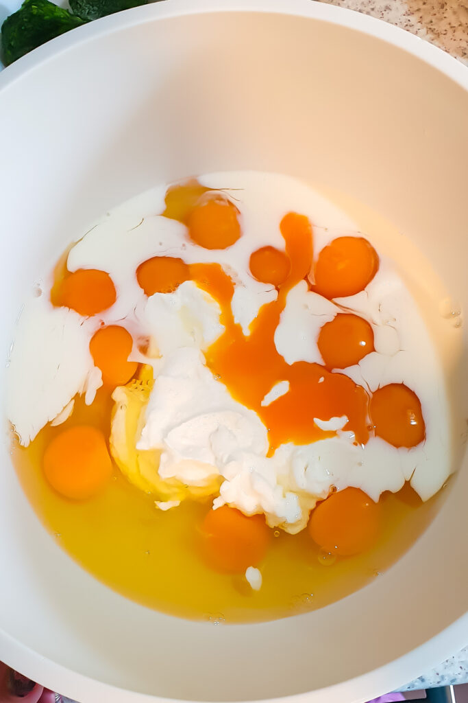 A large white mixing bowl containing several cracked eggs, egg yolks, milk, and a dollop of cottage cheese.