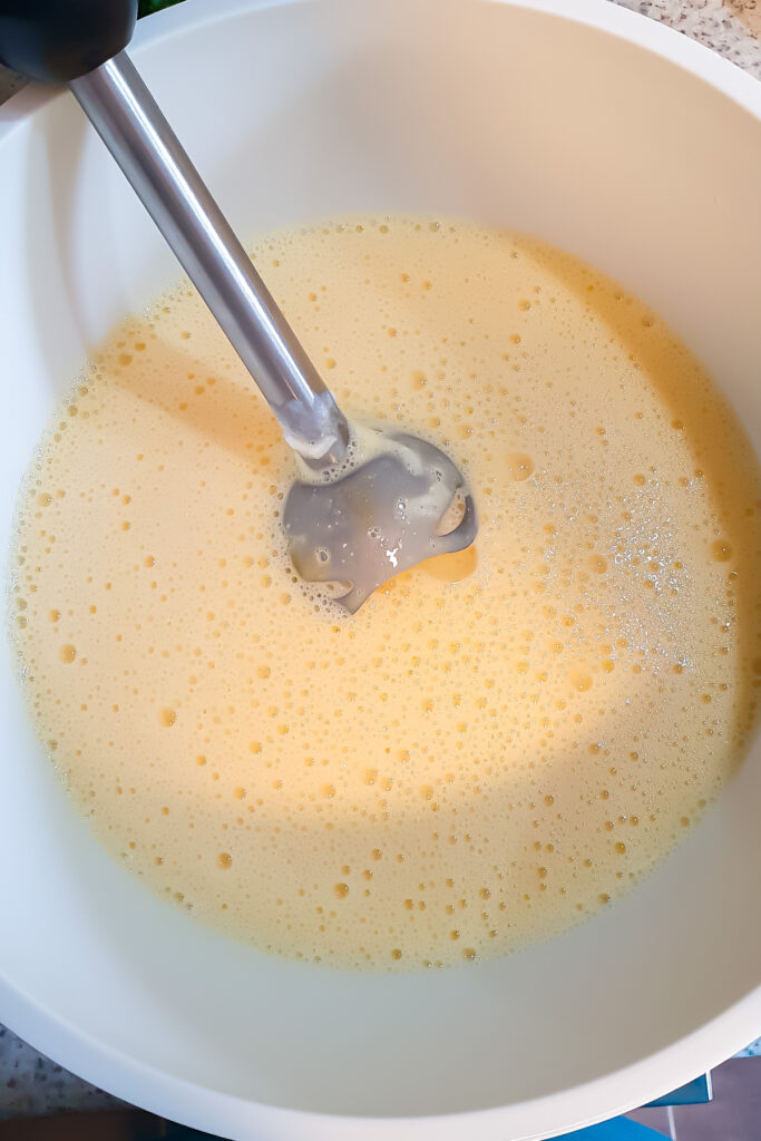 A handheld immersion blender is mixing a light yellow, frothy Cottage Cheese Egg Bake mixture in a large white bowl.