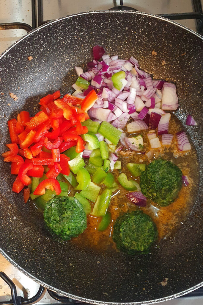 Chopped red onions, red and green bell peppers, and three frozen spinach portions cooking in a black frying pan on a stove.
