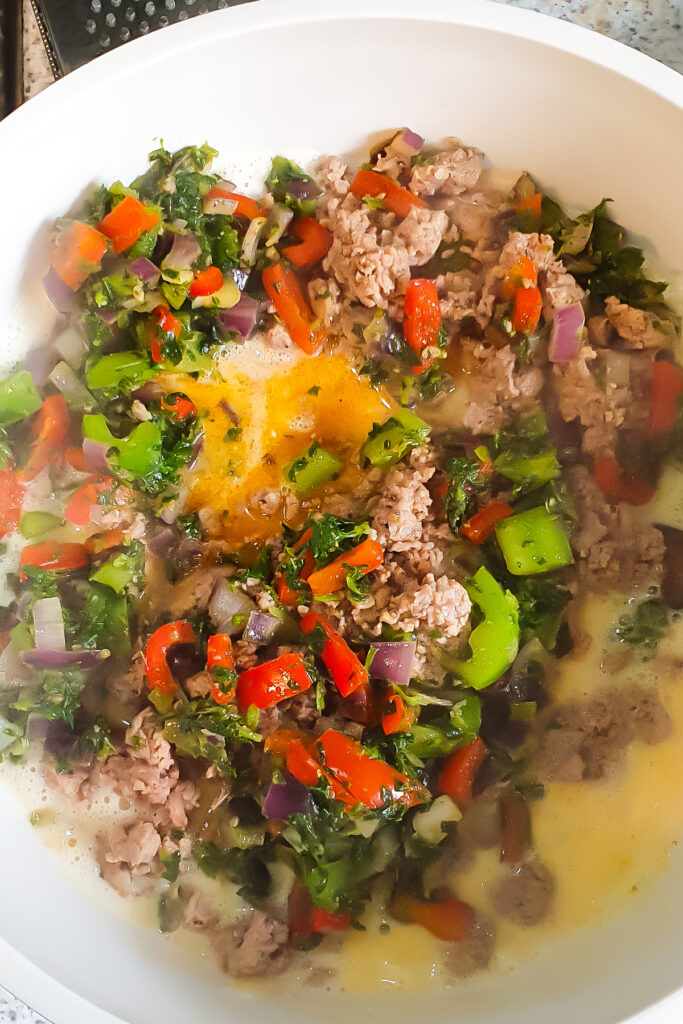 A mixing bowl containing whisked eggs, cottage cheese, chopped red and green bell peppers, red onion, leafy greens, and cooked sausage.