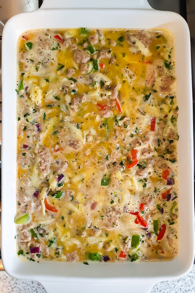 A white baking dish filled with a Cottage Cheese Egg Bake, featuring mixed eggs, chopped vegetables, shredded cheese, cottage cheese, and ground meat, ready to be baked.