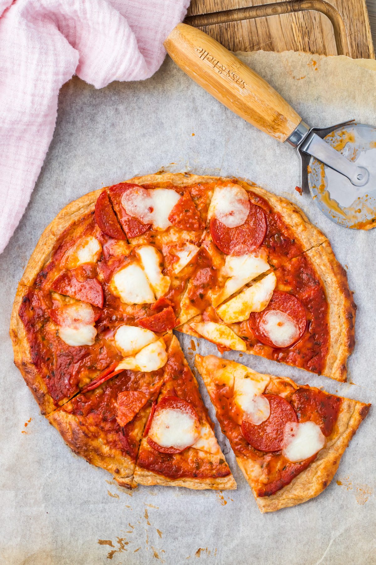 A delicious pepperoni cottage cheese pizza with melted cheese sits on parchment paper, one slice pulled out to tempt you. A pizza cutter and a pink cloth are nearby.
