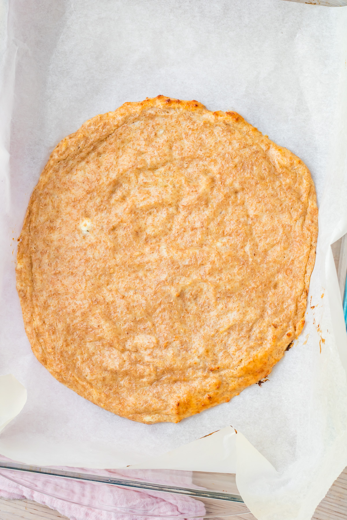 A round, golden-brown cottage cheese pizza crust sits on parchment paper in a glass baking dish.
