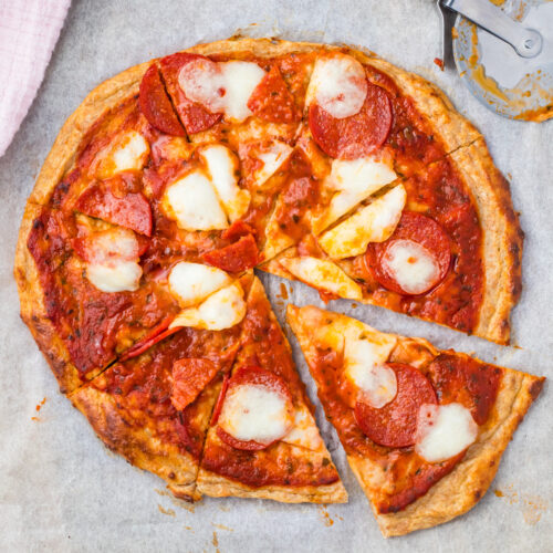 A Cottage Cheese Pizza with melted cheese and pepperoni is cut into eight slices on parchment paper, with a pizza cutter nearby.