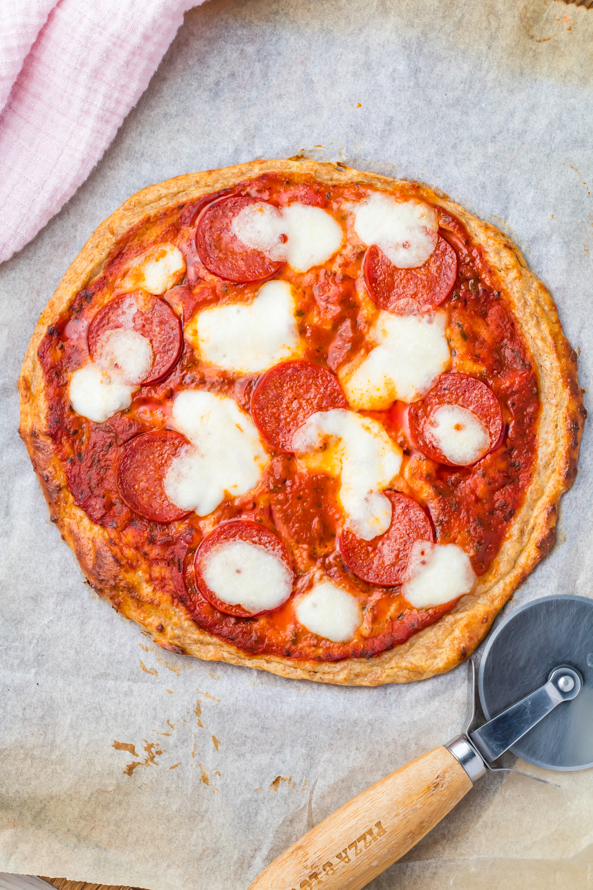 A round high protein cottage cheese pizza with pepperoni and melted cheese on parchment paper, next to a pizza cutter and a pink cloth.