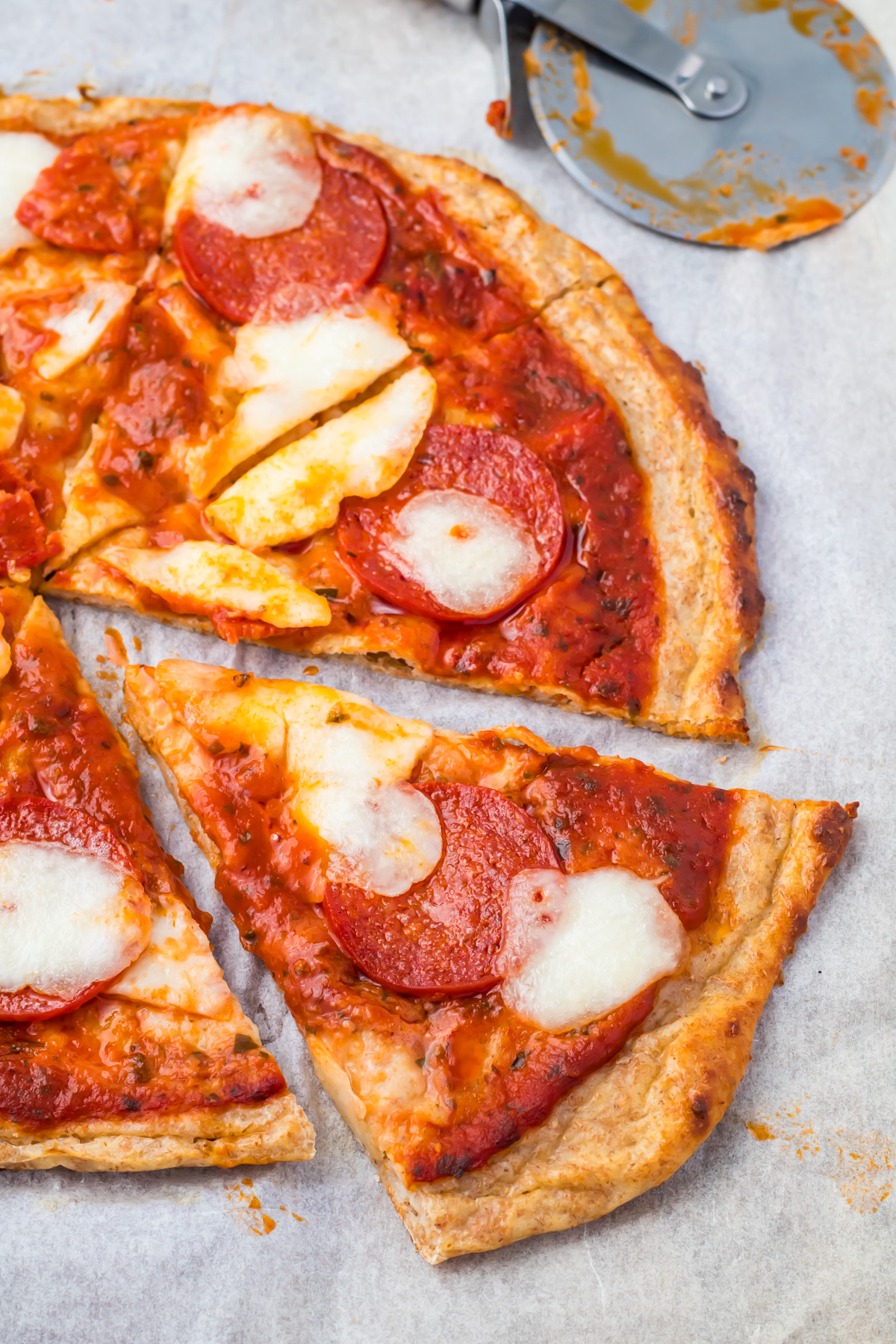 A delicious cottage cheese pizza with pepperoni and slices of mozzarella cheese sits on parchment paper. A pizza cutter lies nearby, and one slice is separated from the rest.