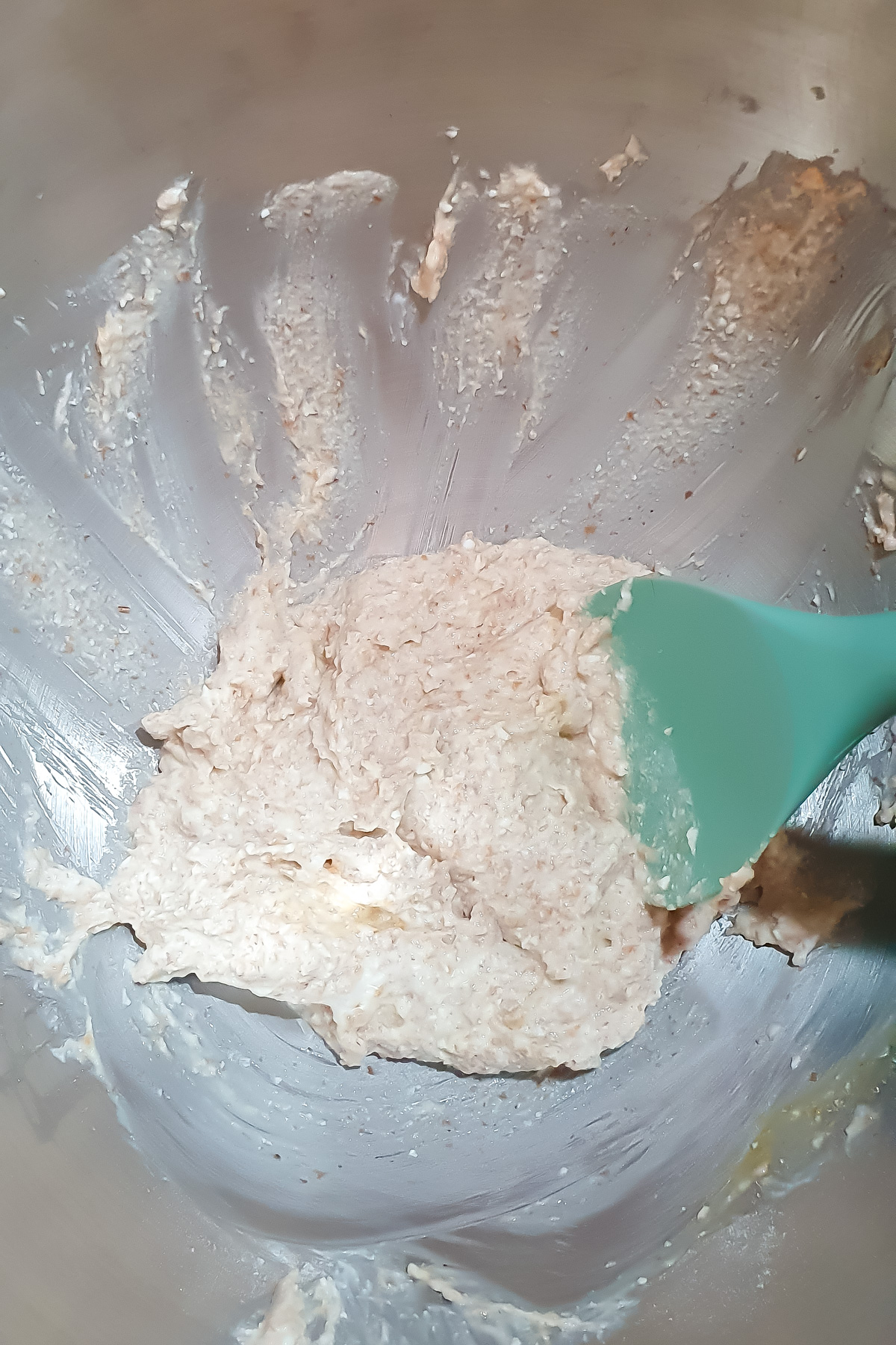 A teal spatula mixing a thick, pale batter or dough in a metal bowl, with some mixture spread along the sides of the bowl.