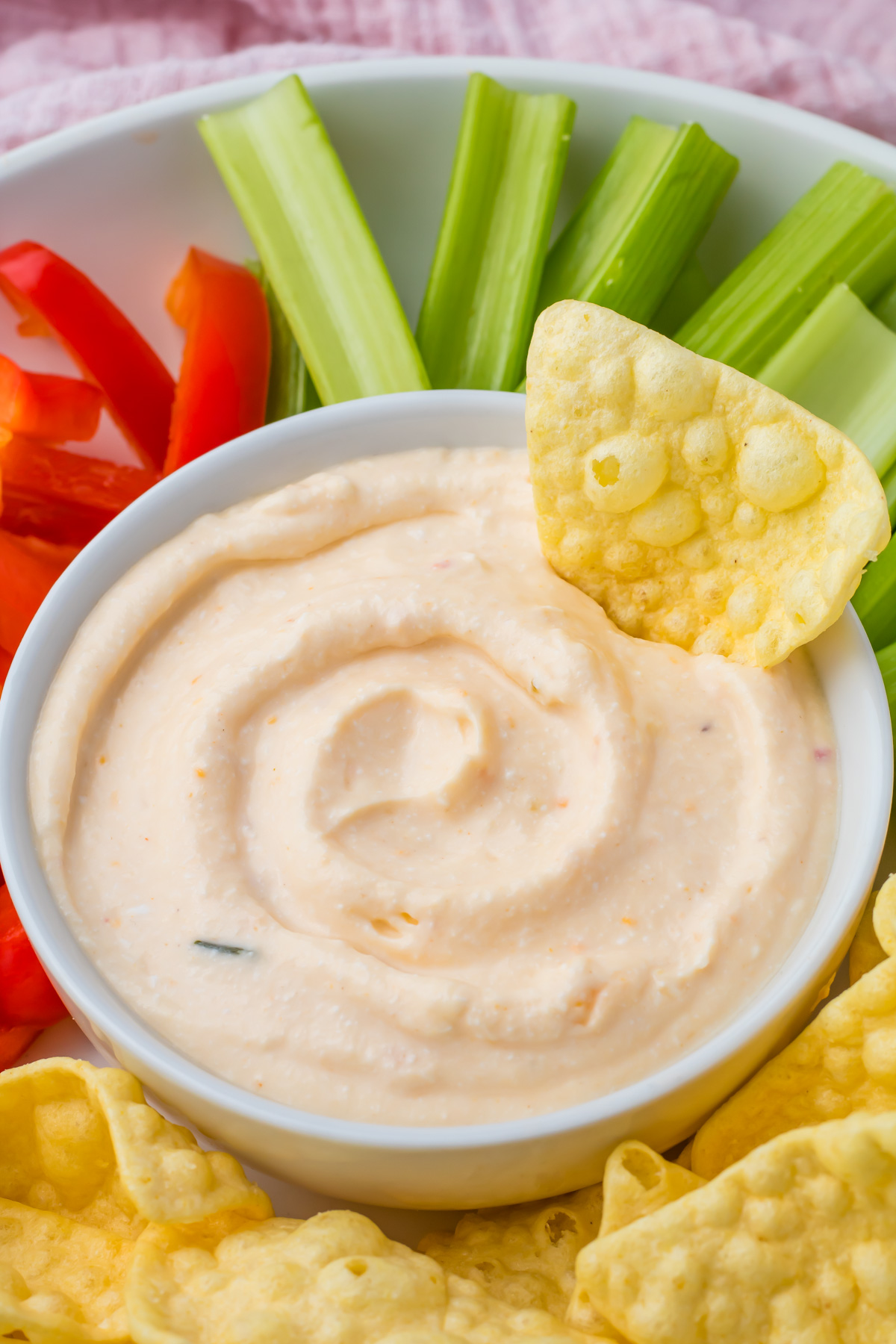 A bowl of creamy Cottage Cheese Queso dip is surrounded by celery sticks, red bell pepper strips, and tortilla chips, with one chip dipped into the bowl.