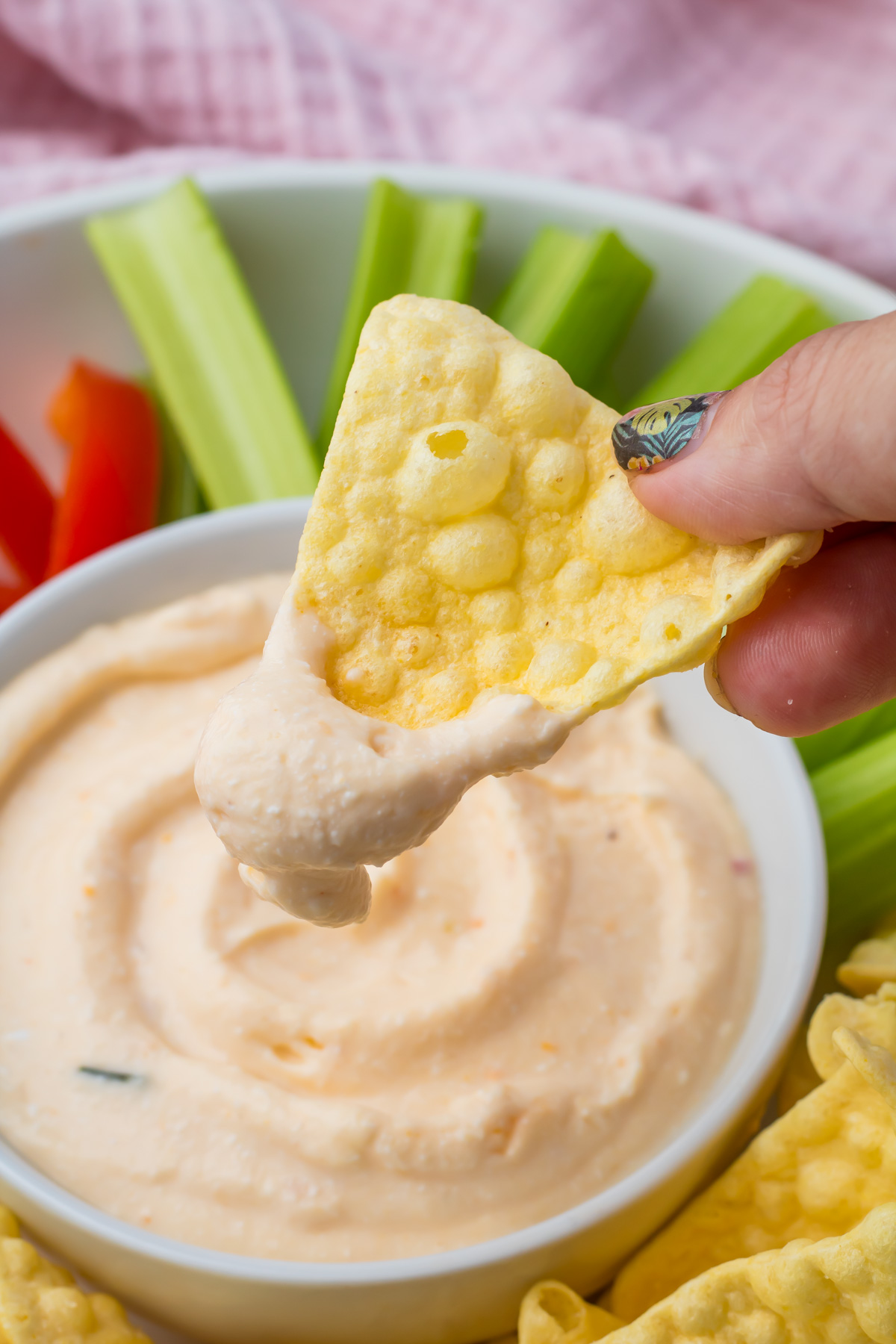 A hand dips a crispy chip into a bowl of creamy Cottage Cheese Queso, surrounded by celery sticks and red bell pepper slices.