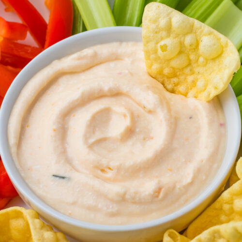 A bowl of creamy Cottage Cheese Queso dip surrounded by celery sticks, red bell pepper slices, and yellow chips, with one chip dipped in the bowl.