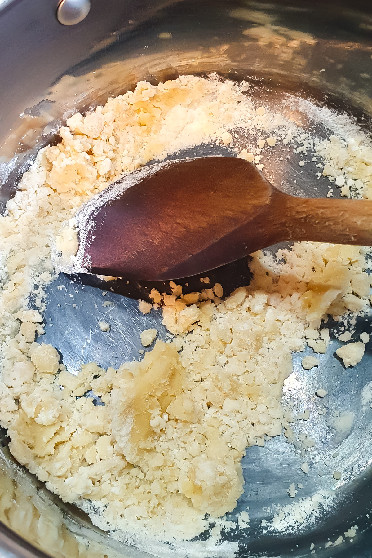 A wooden spoon mixes a clumpy flour and butter mixture in a metal saucepan.
