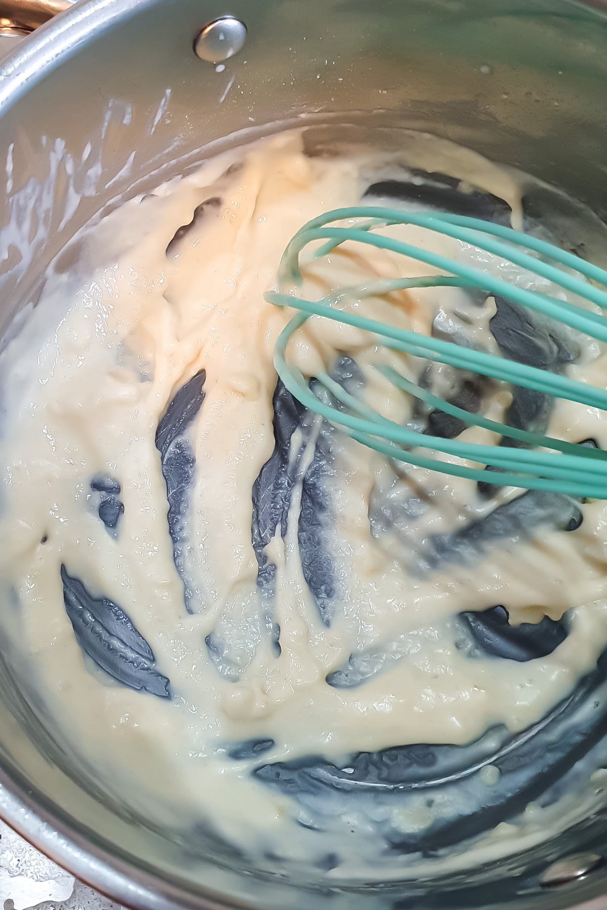 A saucepan containing a thick, creamy white sauce being mixed with a green silicone whisk.