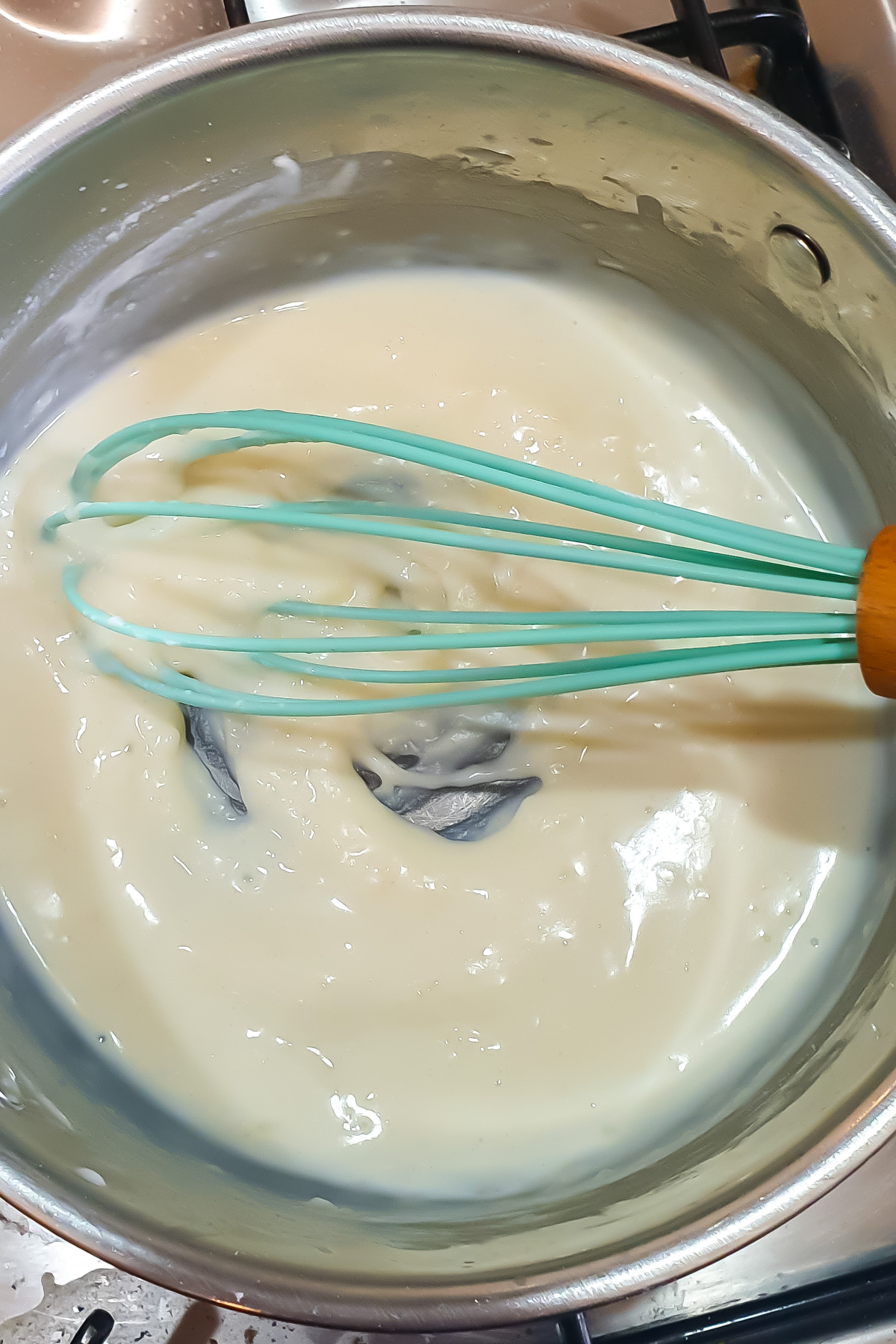 A light blue whisk stirs a thick, creamy roux in a stainless steel pot on a stove.