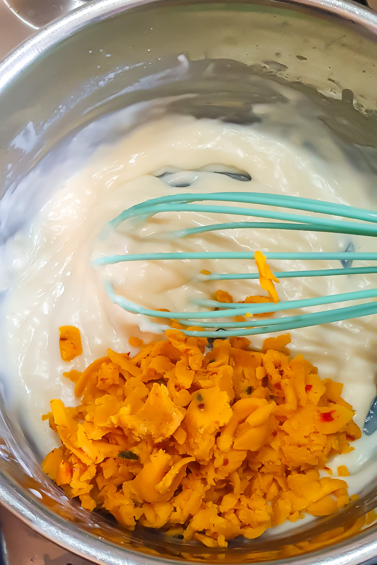 A teal whisk sits in a bowl with a creamy white mixture of and a pile of grated spicy cheese next to it.
