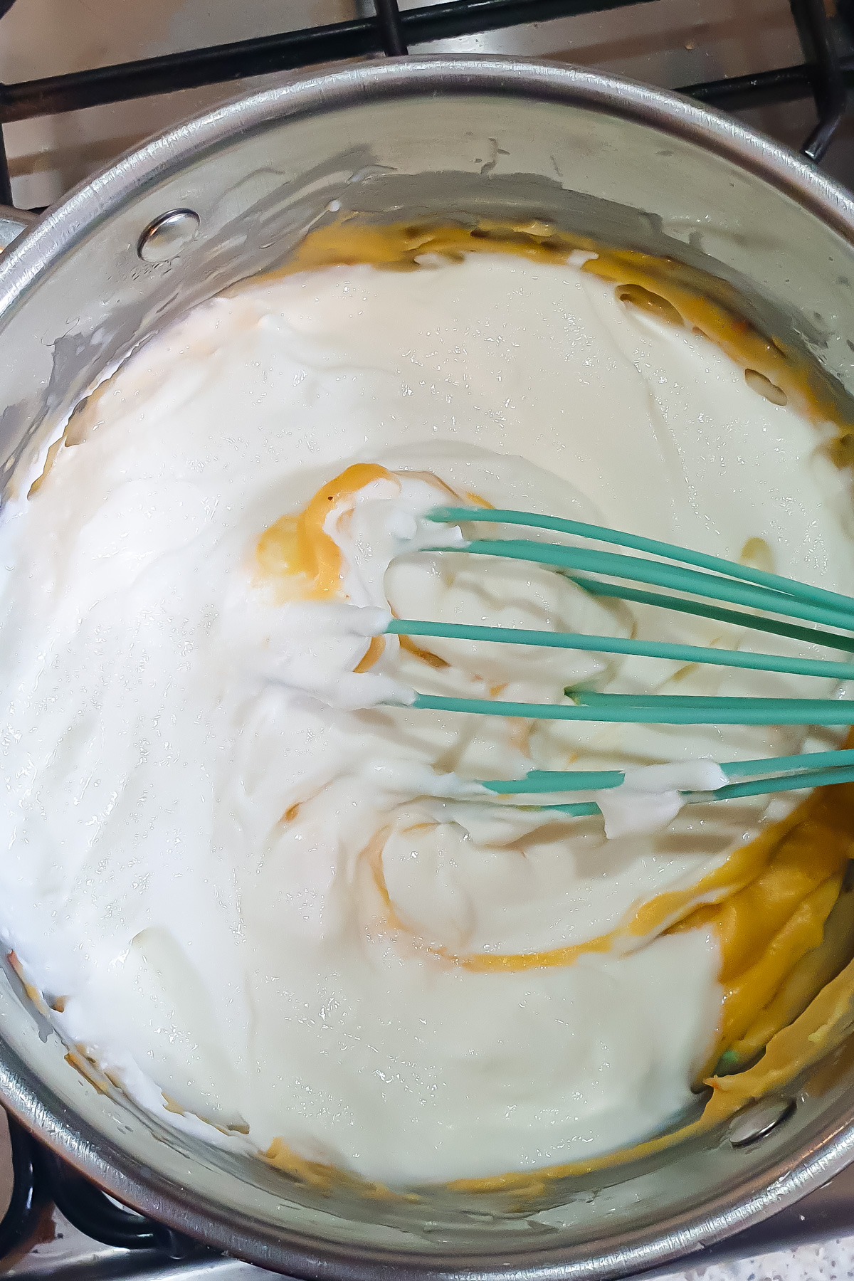 A whisk mixing cottage cheese into a yellow cheesy roux in a metal saucepan on a stove.
