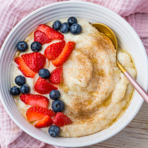 A bowl of creamy egg white oatmeal topped with sliced strawberries, whole blueberries, a sprinkle of cinnamon, and a gold spoon, set on a light pink cloth.