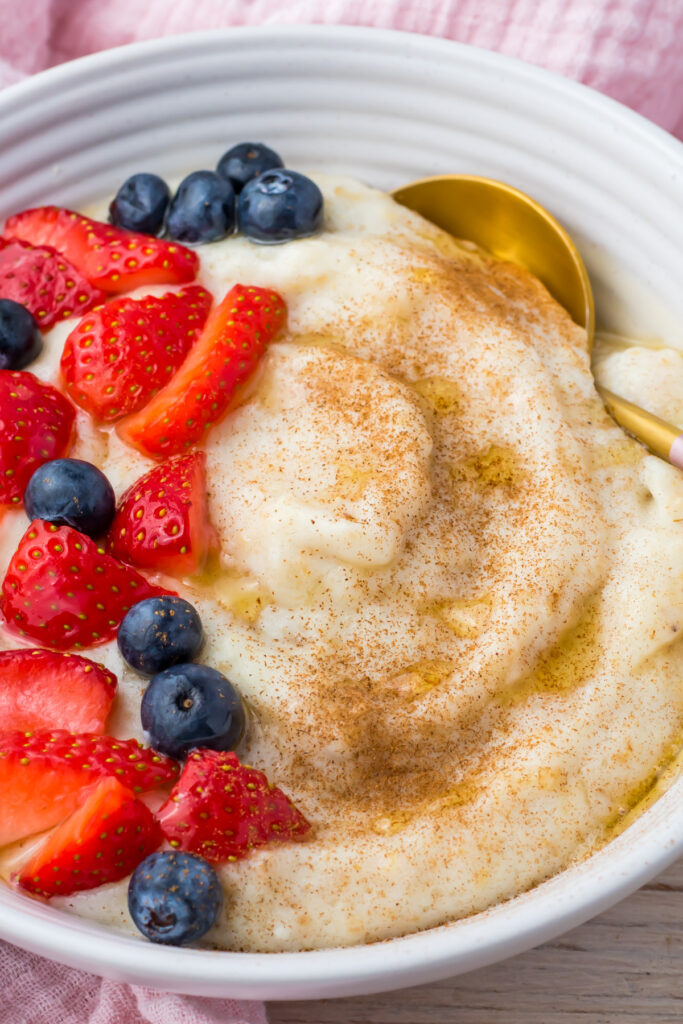 A bowl of creamy egg white oatmeal topped with sliced strawberries, blueberries, a sprinkle of cinnamon, and a drizzle of honey, with a gold spoon on the side.