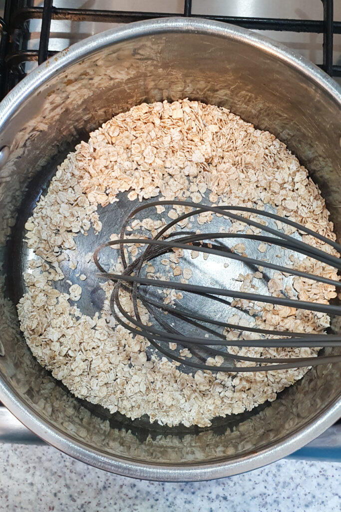 A metal whisk rests on dry rolled oats in a stainless steel saucepan.