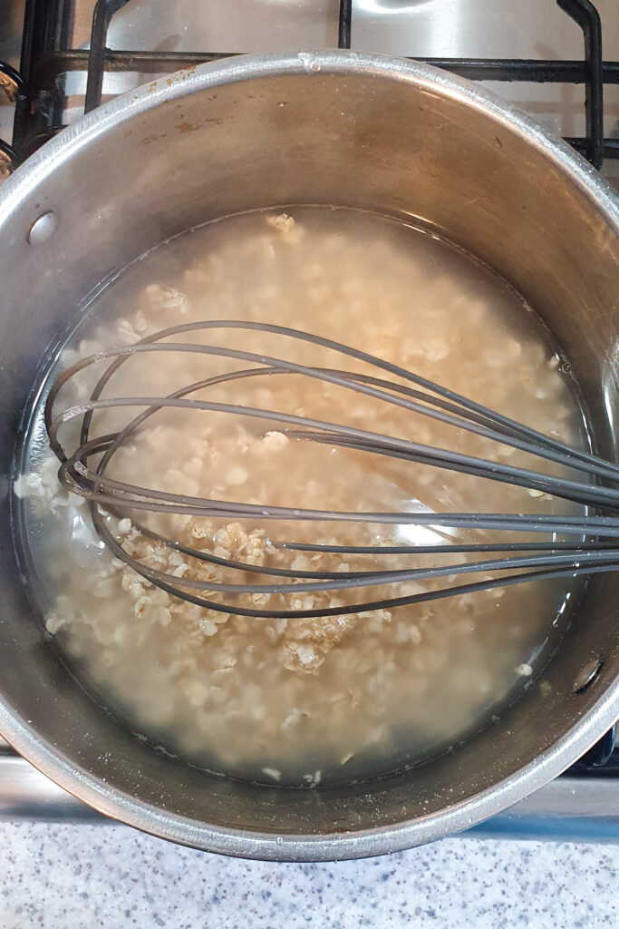 A metal pot containing oats and water is being stirred with a whisk on a stove.