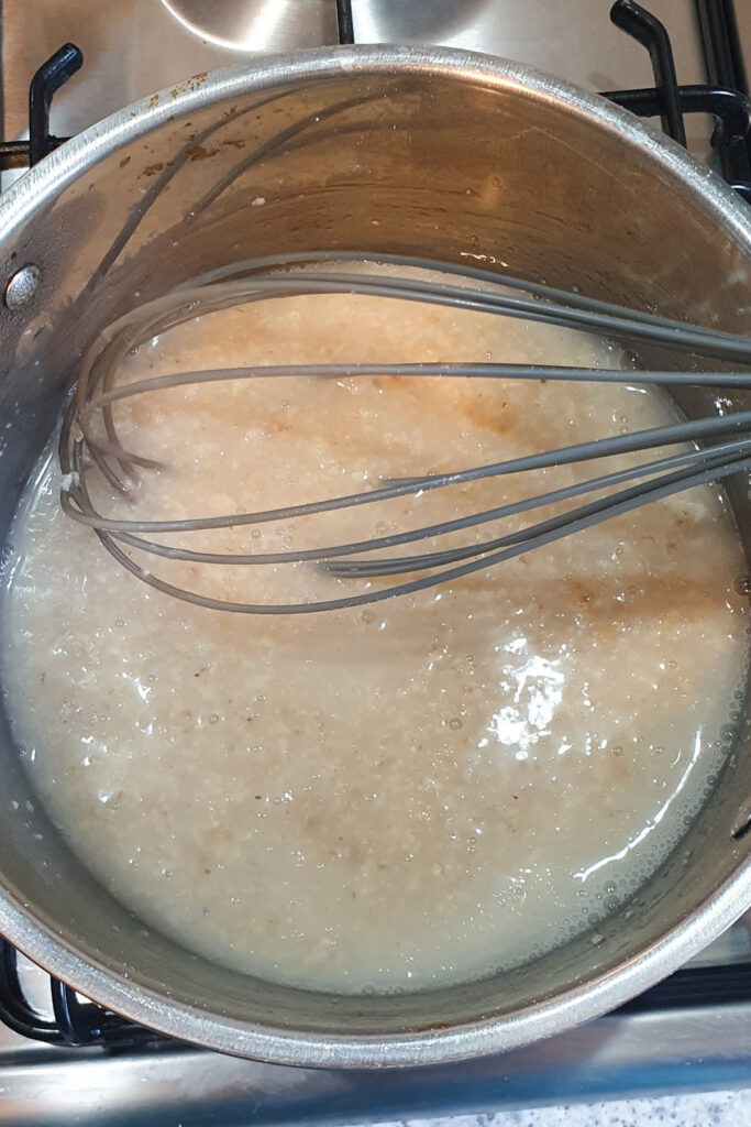 A metal pot on a stove contains partially cooked Oatmeal being stirred with a metal whisk.