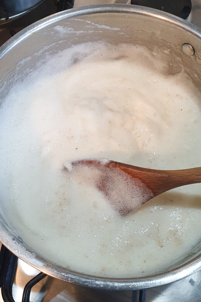 A wooden spoon stirs a pot of bubbling white foamy egg white oatmeal mixture on a stovetop.