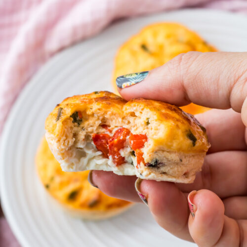 A hand holding a partially eaten starbucks egg white bite filled with cream cheese and red peppers, with more Starbucks egg white bites on a white plate in the background.
