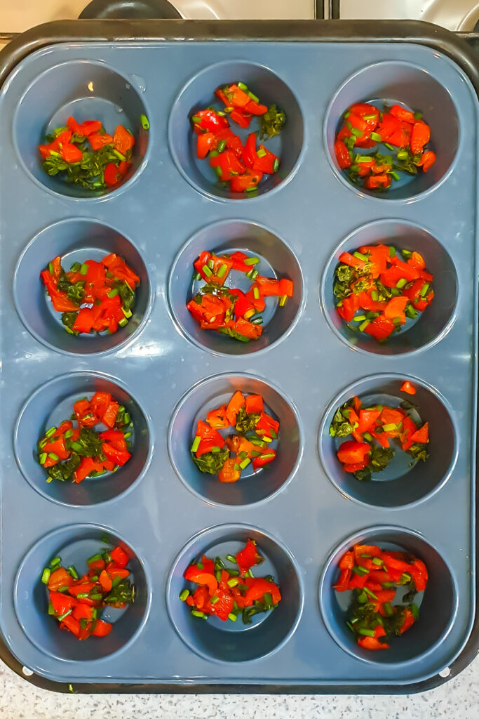 A muffin tray with each cup containing a mixture of chopped red bell peppers and green vegetables, almost ready for baking.