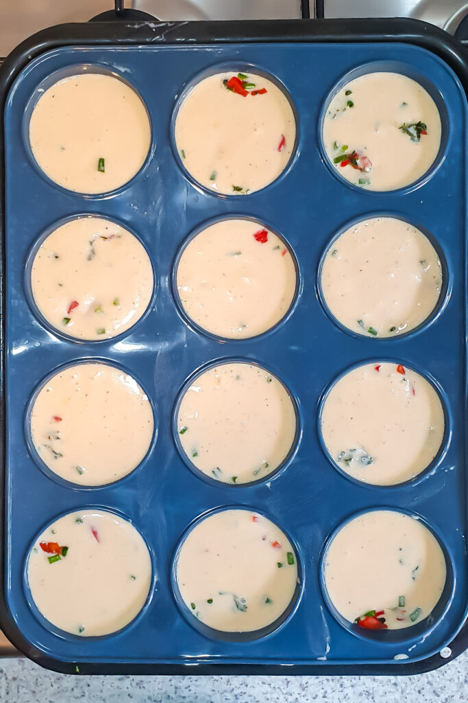 A muffin tin with twelve cups filled with a liquid egg white batter, packed with chopped green herbs and red vegetables, ready to be baked.