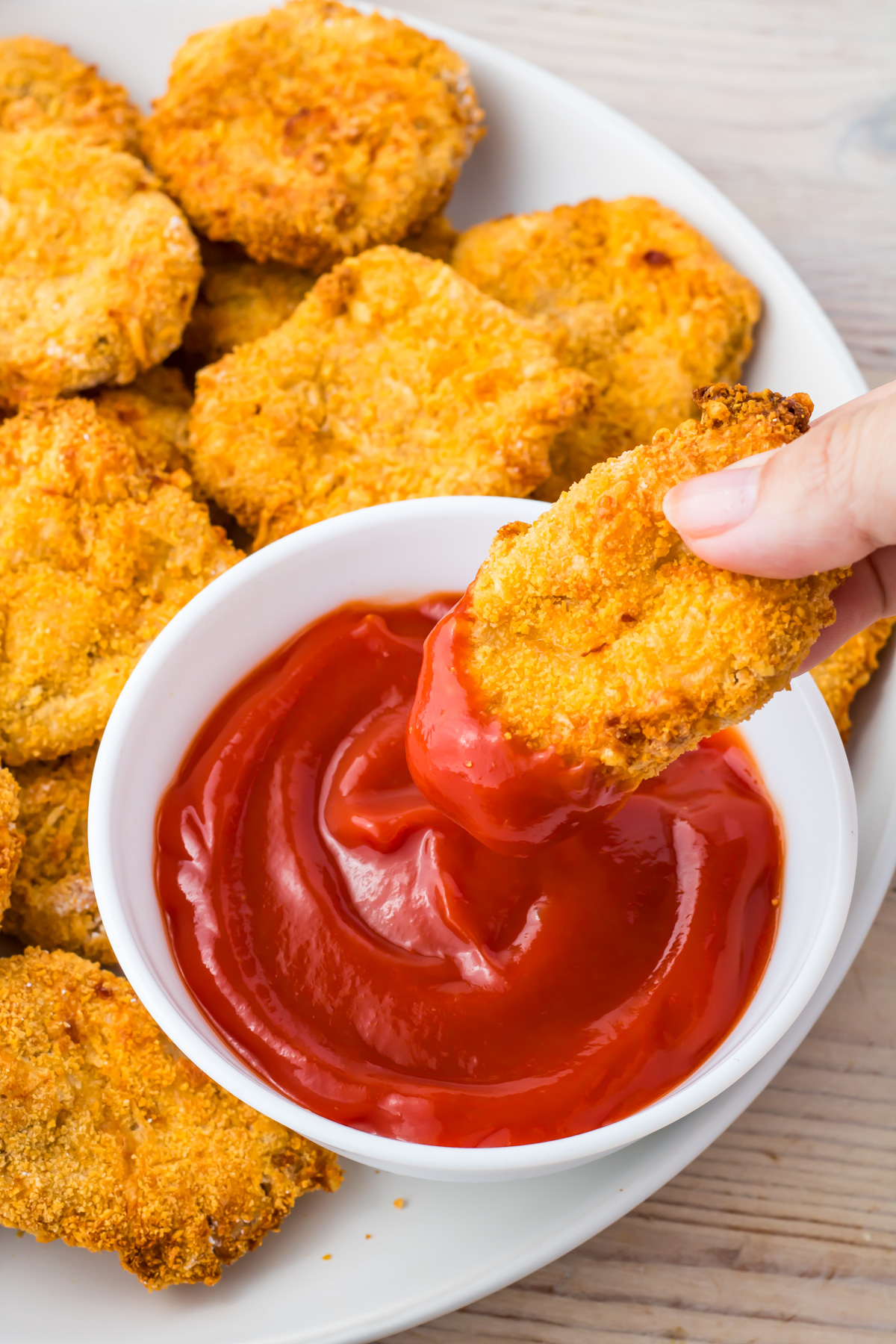 A hand dips a crispy Air Fryer homemade Chicken Nugget into a bowl of ketchup, with more golden nuggets arranged on a white plate in the background.