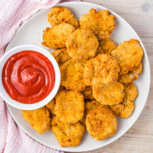 A white plate filled with crispy Air Fryer homemade chicken nuggets made from ground chicken sits next to a small bowl of red dipping sauce, placed on a light wooden surface.