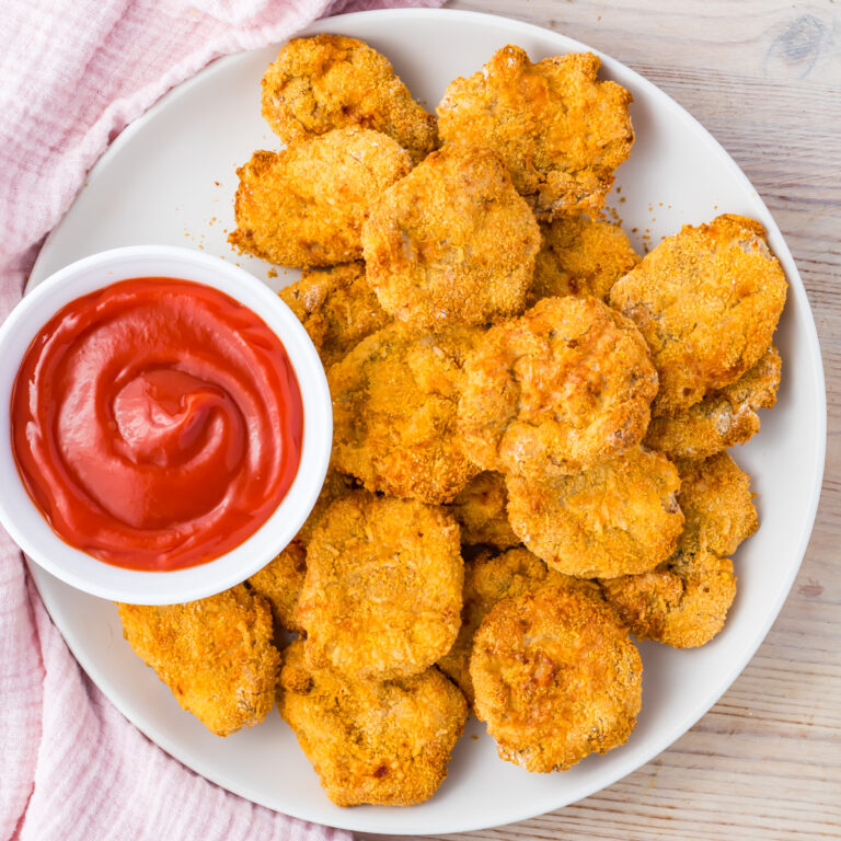A white plate filled with crispy Air Fryer homemade chicken nuggets made from ground chicken sits next to a small bowl of red dipping sauce, placed on a light wooden surface.
