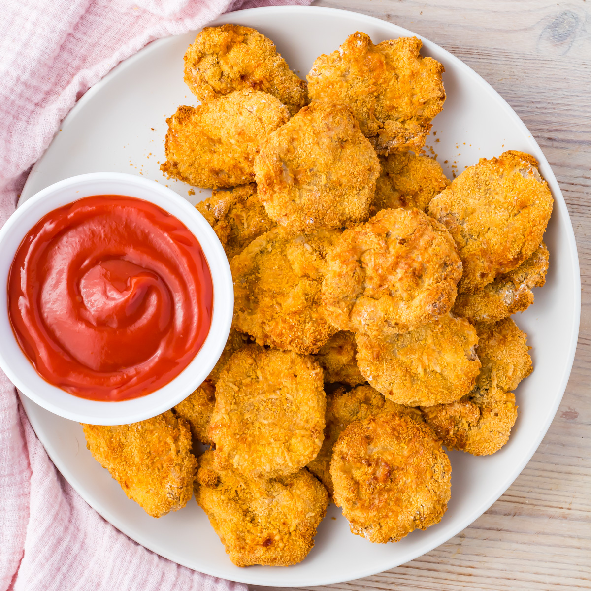 A white plate filled with crispy Air Fryer homemade chicken nuggets made from ground chicken sits next to a small bowl of red dipping sauce, placed on a light wooden surface.
