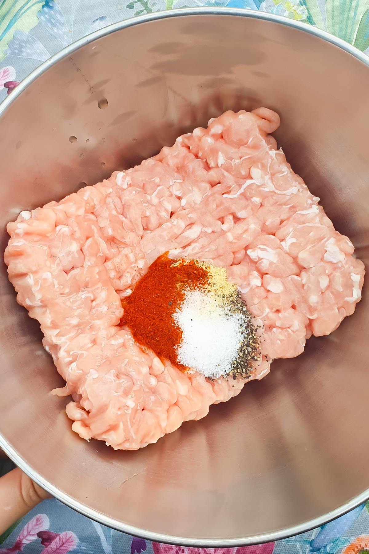 A metal bowl contains raw ground chicken topped with paprika, salt, pepper,  and garlic powder.