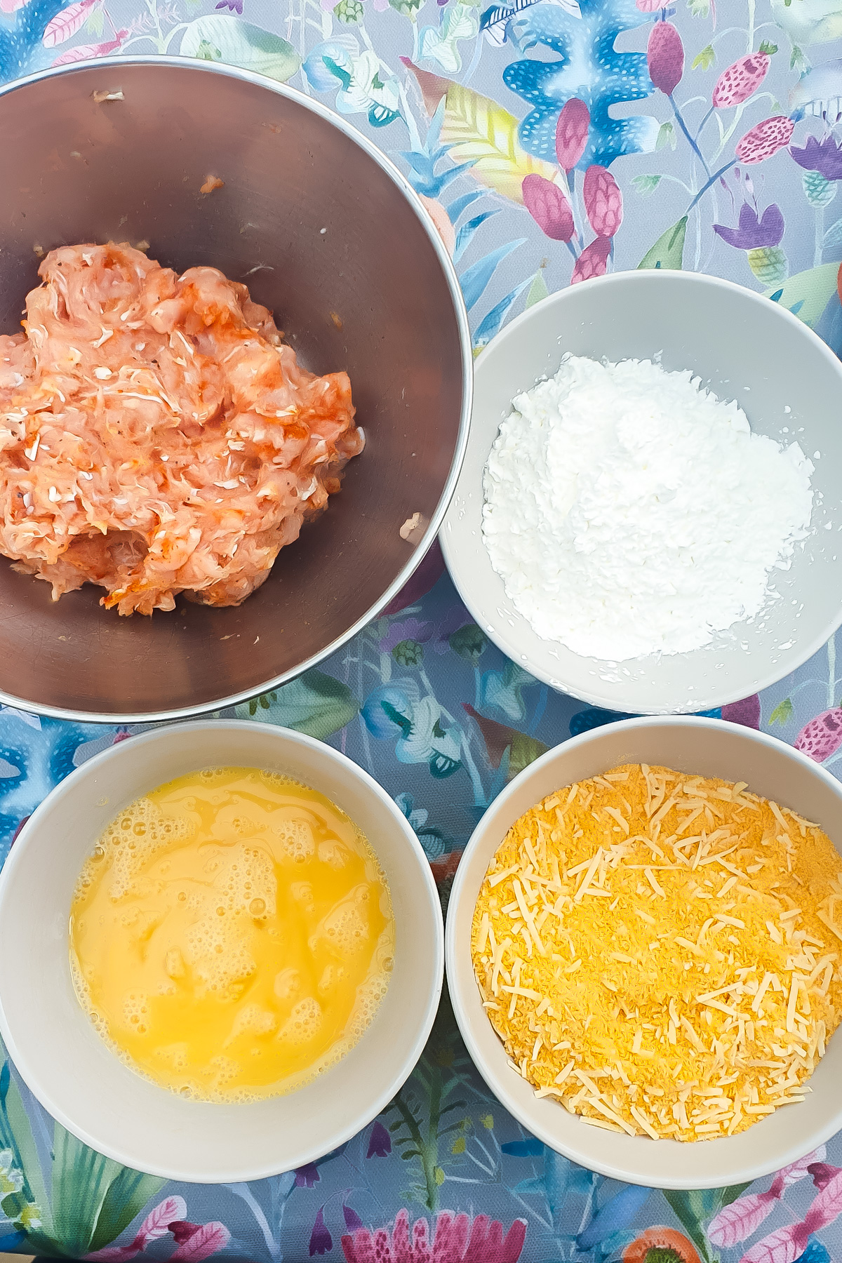 Four bowls on a floral tablecloth hold minced chicken, cornflour, beaten eggs, and breadcrumbs with shredded cheese.