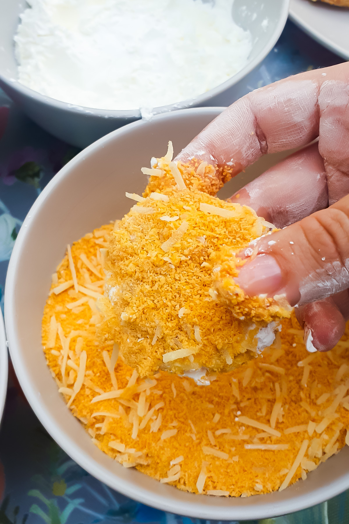 A hand coats a homemade chicken nugget with grated cheese and crumbs in a white bowl.
