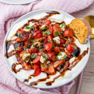 A plate of whipped cottage cheese Bruschetta Dip topped with cherry tomatoes, mozzarella, basil, and balsamic glaze, served with a slice of toasted bread.