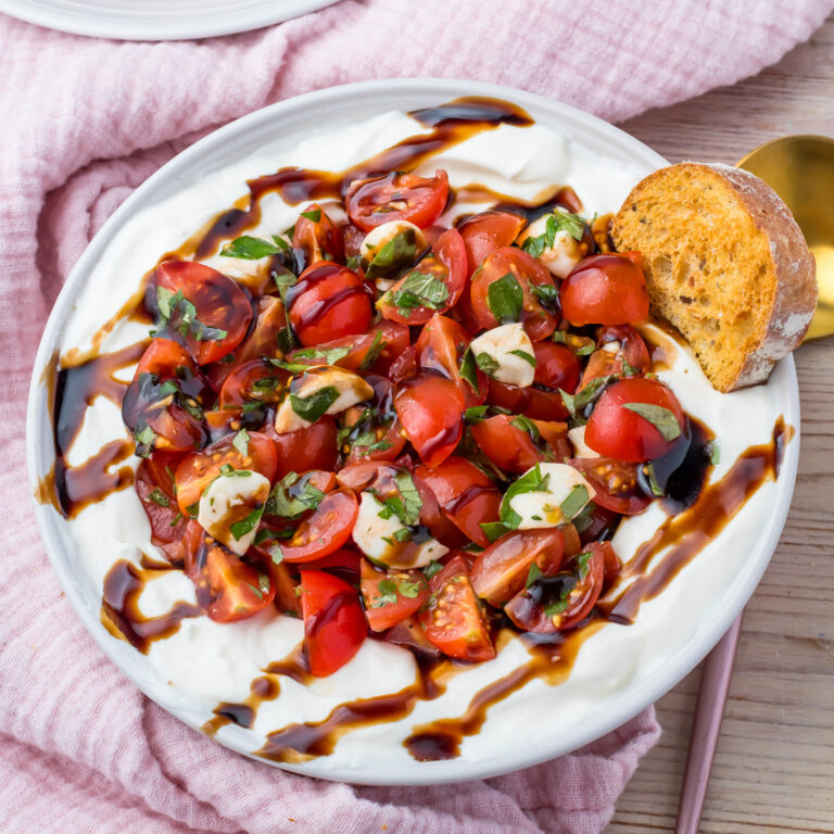 A plate of whipped cottage cheese Bruschetta Dip topped with cherry tomatoes, mozzarella, basil, and balsamic glaze, served with a slice of toasted bread.