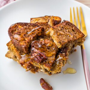 A serving of high protein French toast casserole with pecans and drizzled syrup on a white plate with a gold fork.