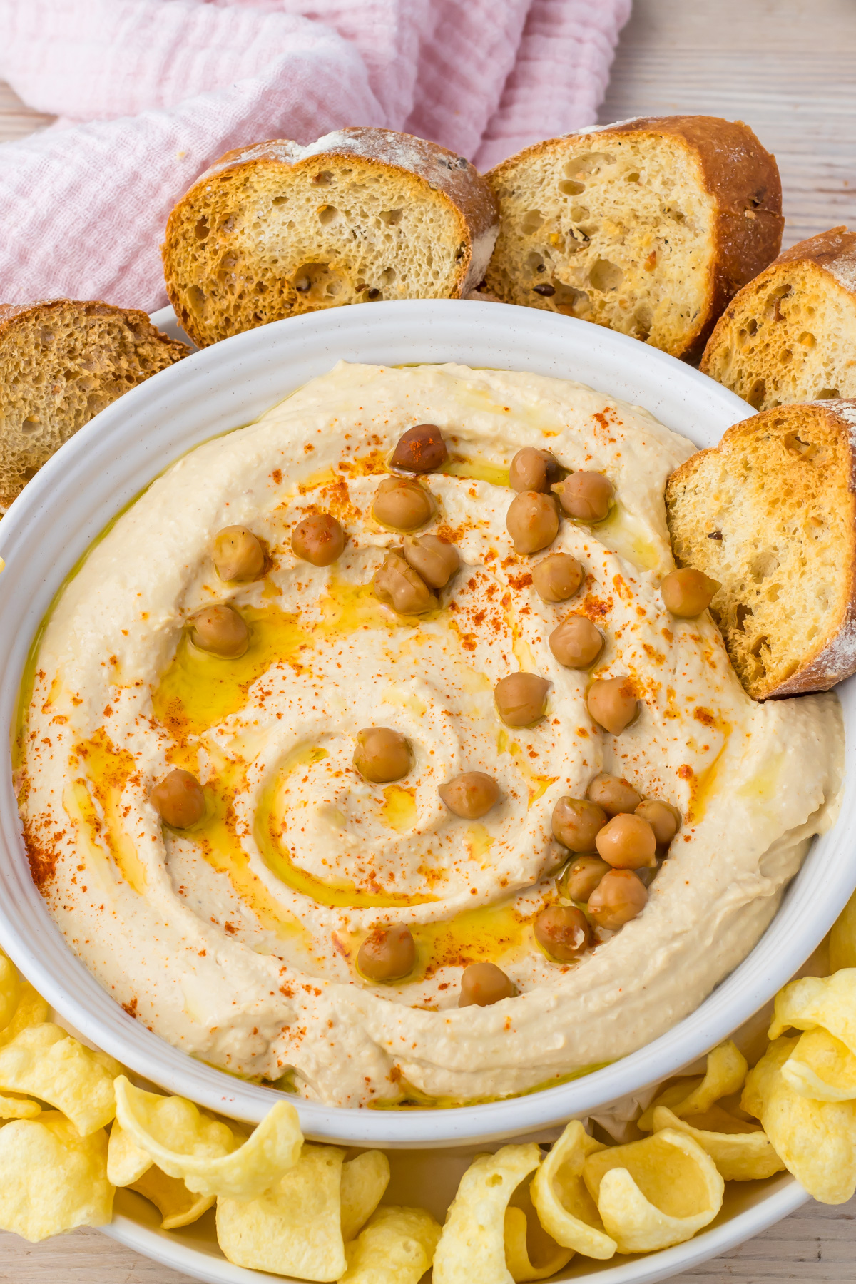 A bowl of high protein hummus topped with whole chickpeas, paprika, and olive oil, surrounded by slices of bread and potato chips.