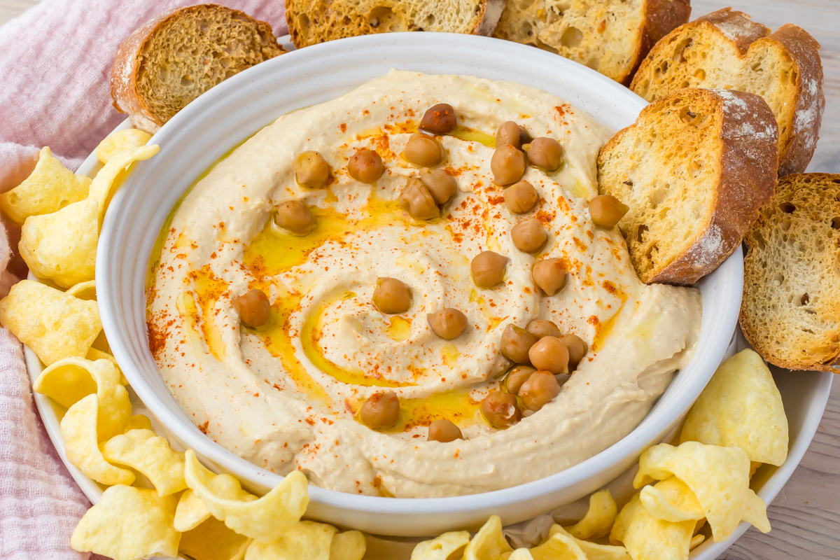 A bowl of high protein hummus topped with whole chickpeas, olive oil, and paprika, surrounded by slices of toasted bread and yellow chips.