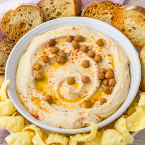 A bowl of high protein hummus topped with whole chickpeas, olive oil, and paprika, surrounded by toasted bread slices and chips.