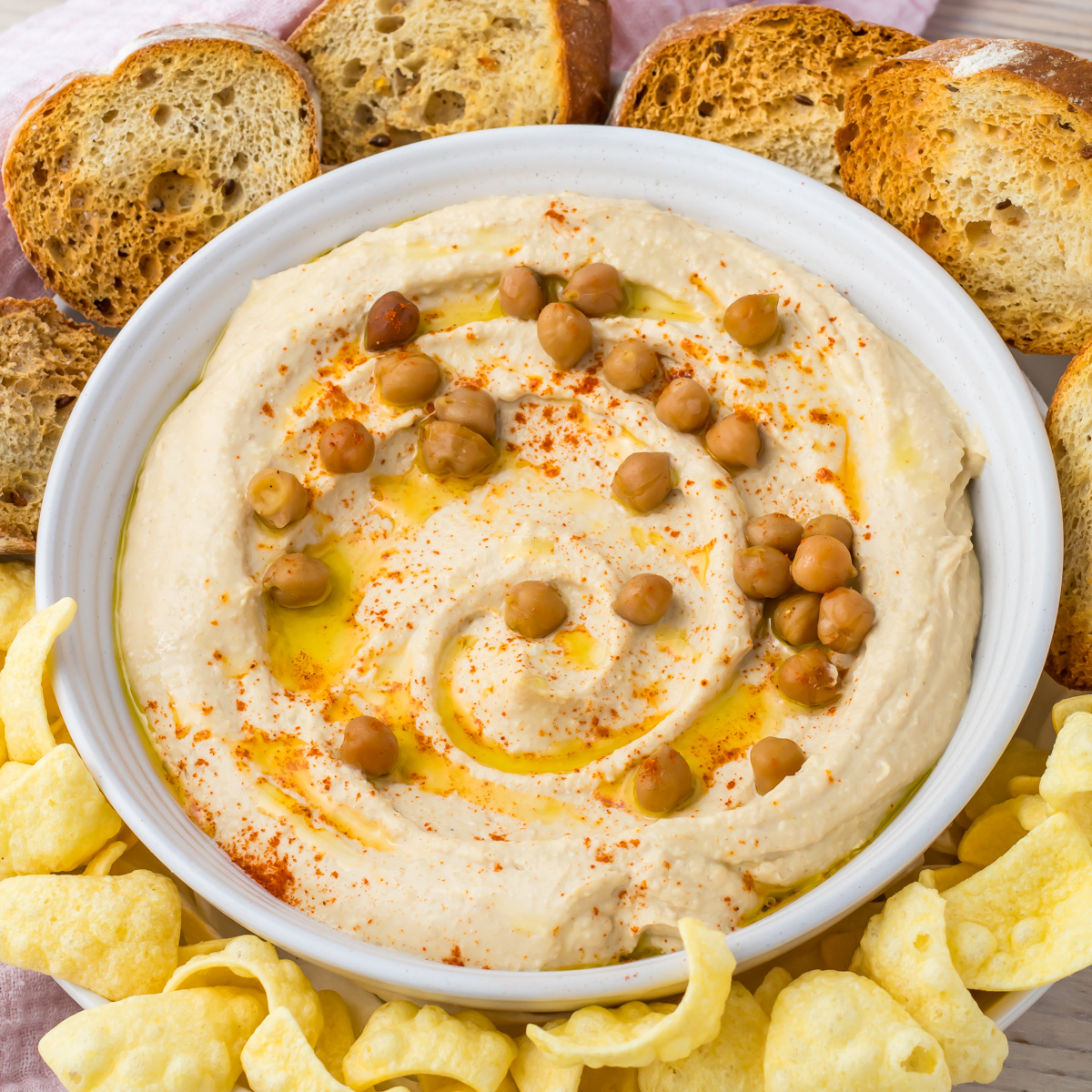 A bowl of high protein hummus topped with whole chickpeas, olive oil, and paprika, surrounded by toasted bread slices and chips.
