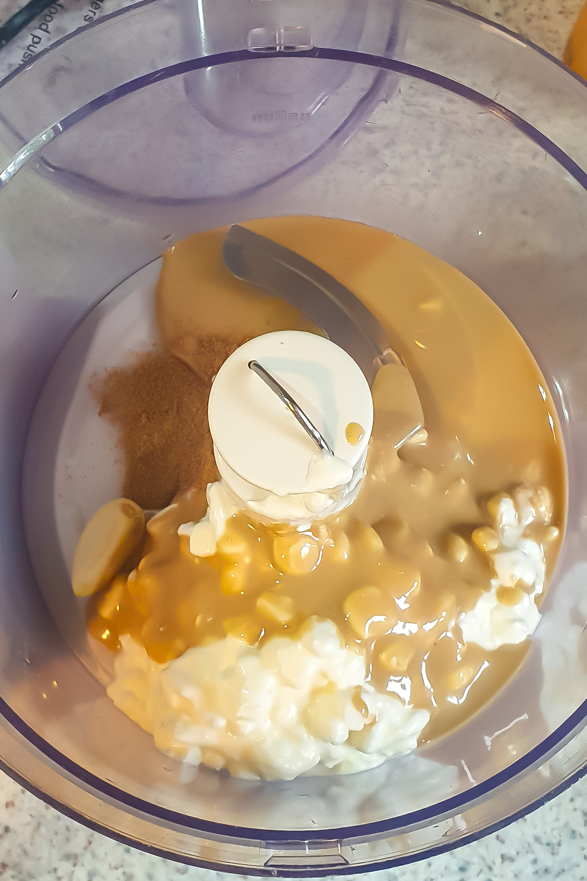 A food processor bowl containing cottage cheese, tahini, cumin powder, and garlic is ready to be blended.