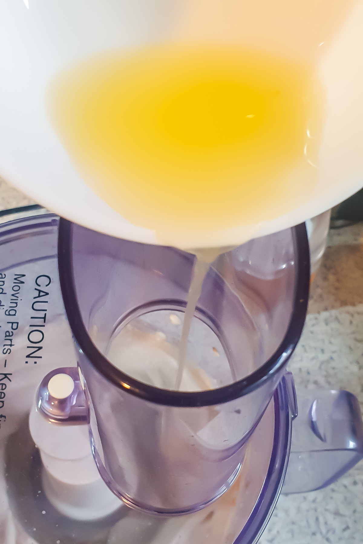Lemon juice is being poured from a white bowl into the chute of a food processor on a kitchen counter.