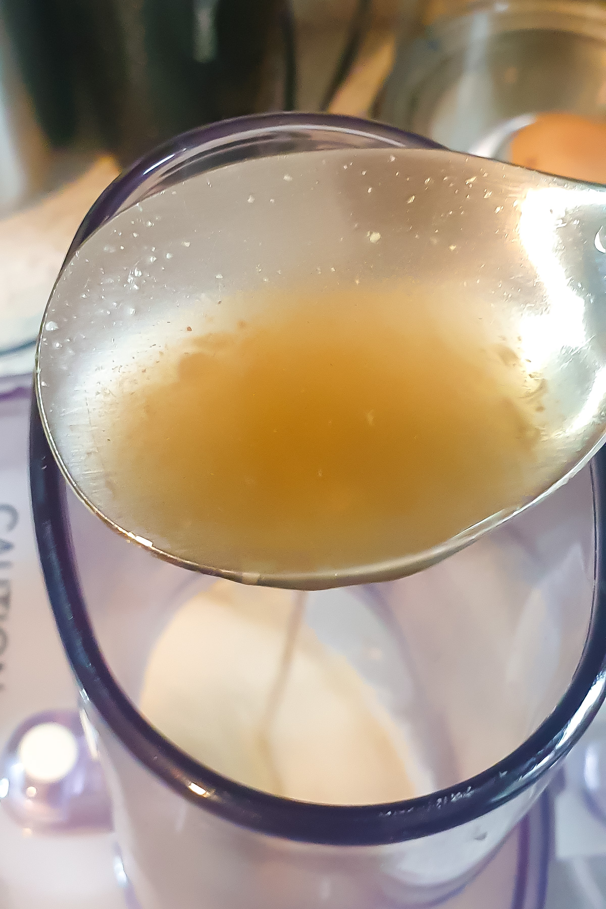 A close-up of a spoon holding a small amount of aquafaba being drizzled into a food processor.