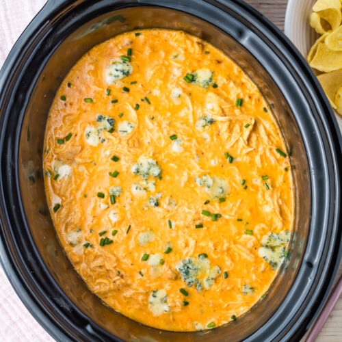 Slow cooker filled with creamy Buffalo Chicken Dip, garnished with blue cheese crumbles and chopped chives, with tortilla chips on a plate nearby.