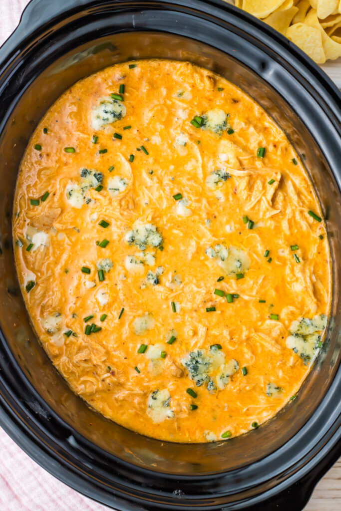 Slow Cooker filled with creamy Buffalo Chicken Dip, garnished with chopped chives and blue cheese crumbles. Chips are partially visible in the background.