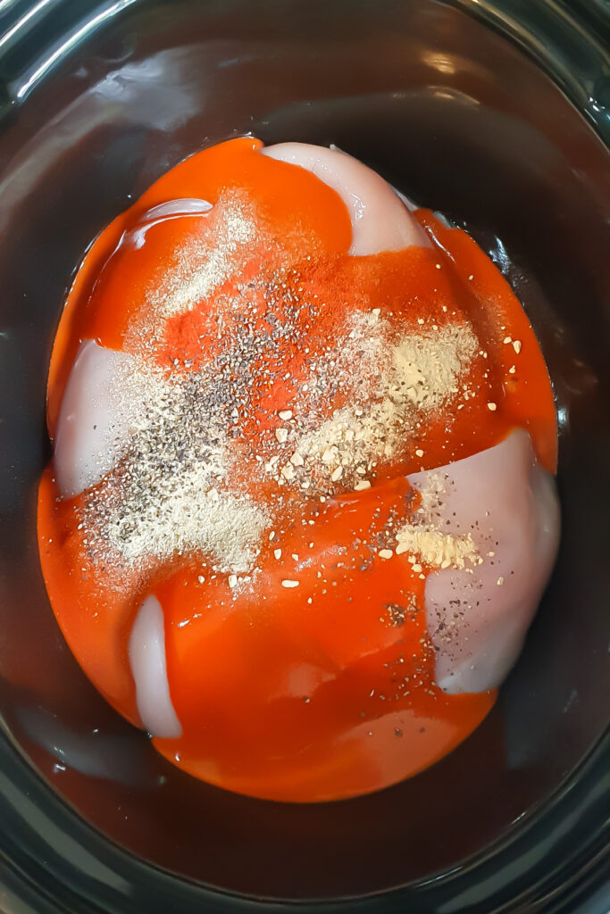 Raw chicken breasts in a slow cooker, topped with red sauce and a blend of black pepper, garlic powder, and other seasonings.