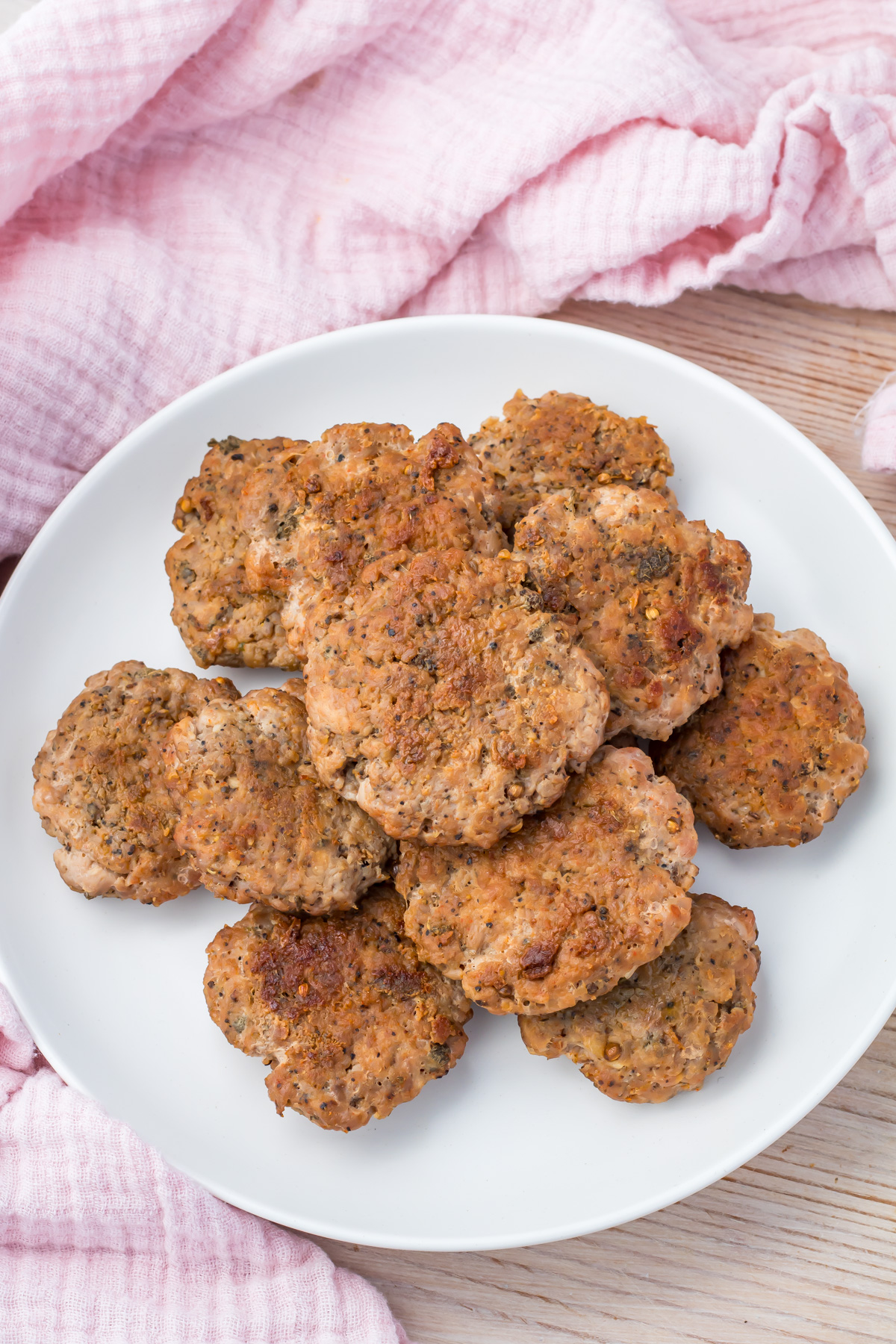 A white plate with several browned turkey breakfast sausage patties, placed on a light wooden surface with a pink cloth in the background.