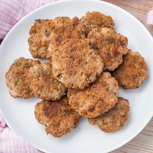 A white plate filled with several cooked, seasoned turkey breakfast sausage patties sits on a light wooden surface.